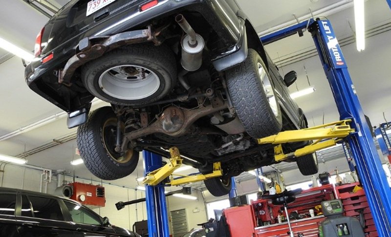 Car raised on a hydraulic lift in an auto repair shop, viewed from underneath, showing the exhaust, rear axle, suspension, and tires with blue and yellow lift arms overhead.