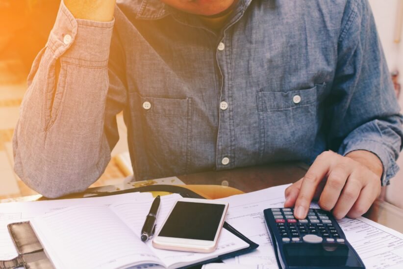 Person in a denim shirt calculating finances at a desk with a calculator, smartphone, and notebook open in front of them