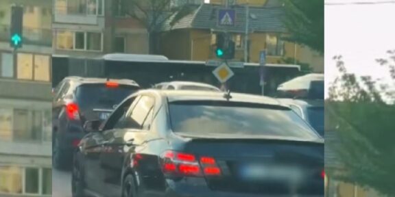 Rear view of a dark sedan in a traffic jam on a city street with apartment buildings and trees in the background.