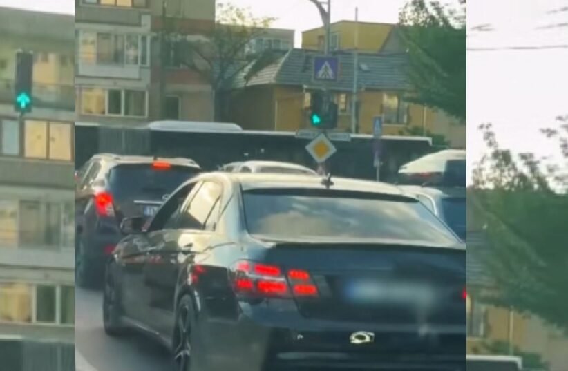 Rear view of a dark sedan in a traffic jam on a city street with apartment buildings and trees in the background.