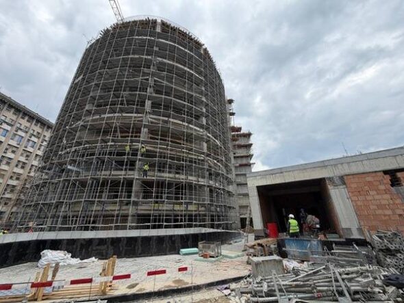 Cylindrical building under heavy scaffolding at an active construction site, with workers near a tunnel-like entrance nearby.