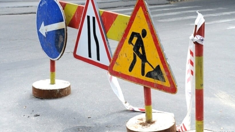 Road work zone with warning signs and barriers on an asphalt street; includes a blue left-turn sign, lane merge sign, and digging worker symbol with tape barriers