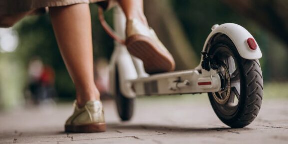 Person wearing beige pants and sneakers stands next to a white kick scooter on a paved path, viewed low from the ground.