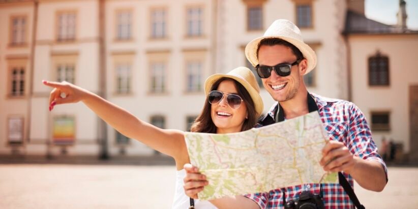Couple wearing sun hats and sunglasses looking at a map while exploring a sunny city square; one points into the distance.