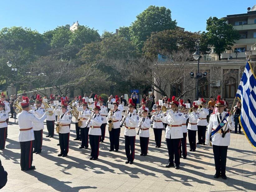 Military band in white uniforms performing a parade in a sunlit square, with a Greek flag on the right.