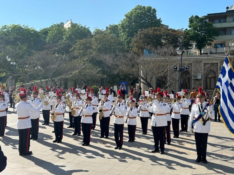 Military band in white uniforms performing a parade in a sunlit square, with a Greek flag on the right.