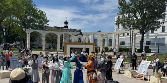 Group of performers in vintage costumes posing and dancing in a sunny outdoor plaza; audience watches in the background.
