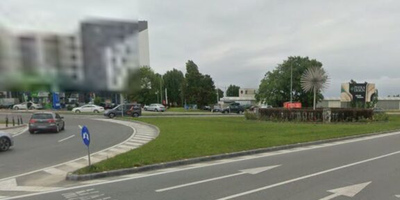 Roundabout with a grassy median, several cars, and a spiky spherical sculpture in the center; buildings in the background under a cloudy sky.