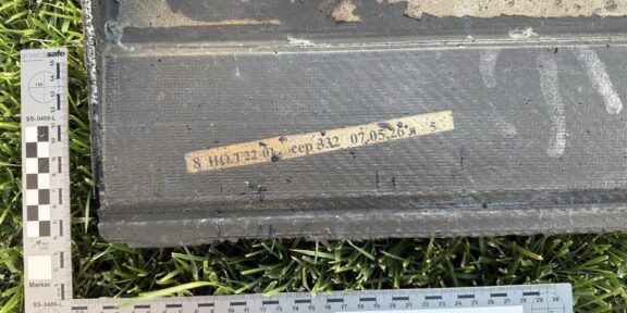 Black rectangular case on grass with a beige labeled tape and two measurement rulers nearby, suggesting field evidence tagging.