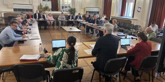 Large formal meeting in a U-shaped wooden-floored chamber with attendees seated around tables, some using laptops and papers on display.