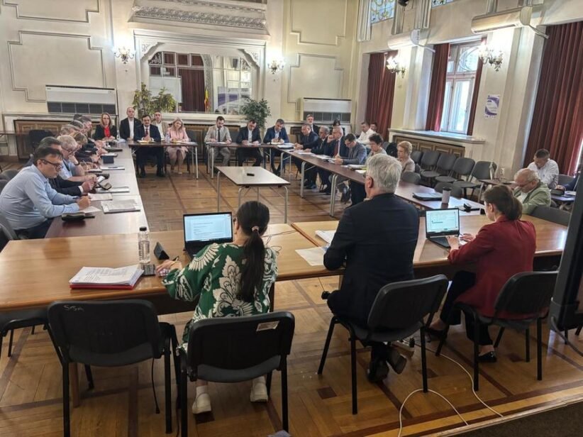 Large formal meeting in a U-shaped wooden-floored chamber with attendees seated around tables, some using laptops and papers on display.