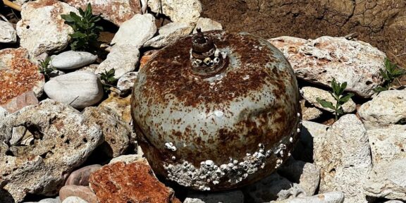 Rusty spherical buoy covered with barnacles resting among rocks on shore