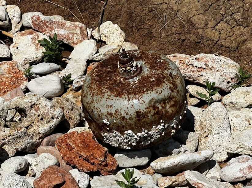 Rusty spherical buoy covered with barnacles resting among rocks on shore