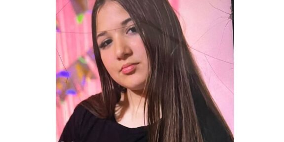 Portrait of a young woman with long dark hair wearing a black top, posed against a pink backdrop with soft makeup.