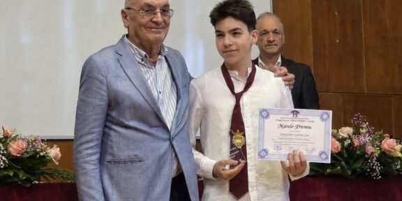 Student in a white shirt and burgundy tie holds a certificate and gold medal, flanked by two men at an awards ceremony on stage with flowers nearby.