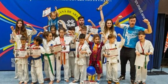 Group of children in karate gis posing with certificates and medals on a gym stage, with a coach on the right and a blue banner behind them.