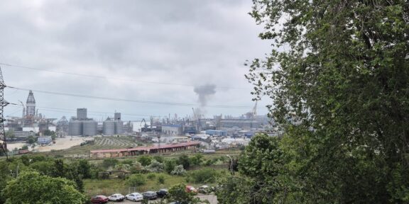 Industrial port scene with cranes, silos and a smokestack, viewed from a green hillside under a cloudy sky.