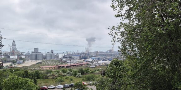 Industrial port scene with cranes, silos and a smokestack, viewed from a green hillside under a cloudy sky.