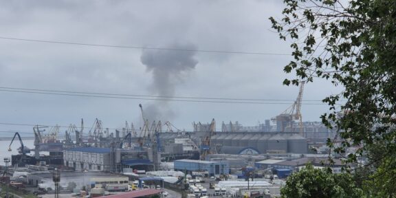 Industrial port scene with cranes and warehouses; a dark smoke plume rising into cloudy sky in the distance