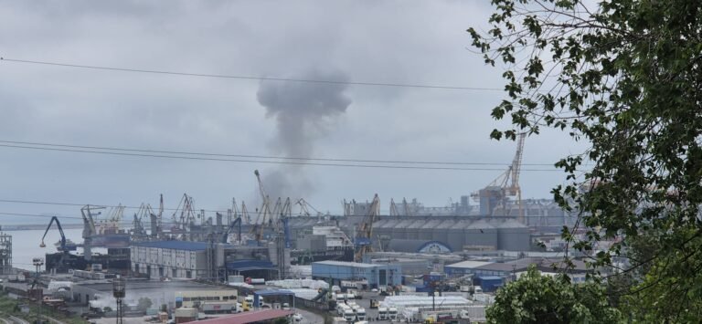 Industrial port scene with cranes and warehouses; a dark smoke plume rising into cloudy sky in the distance
