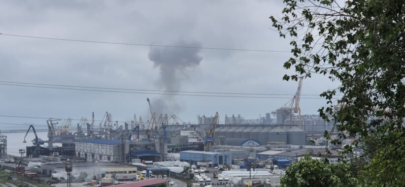 Industrial port scene with cranes and warehouses; a dark smoke plume rising into cloudy sky in the distance