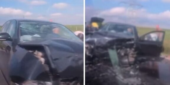 Black sedan with heavily damaged front end after a crash; airbags deployed and driver-side door open on a rural highway with debris on the road.