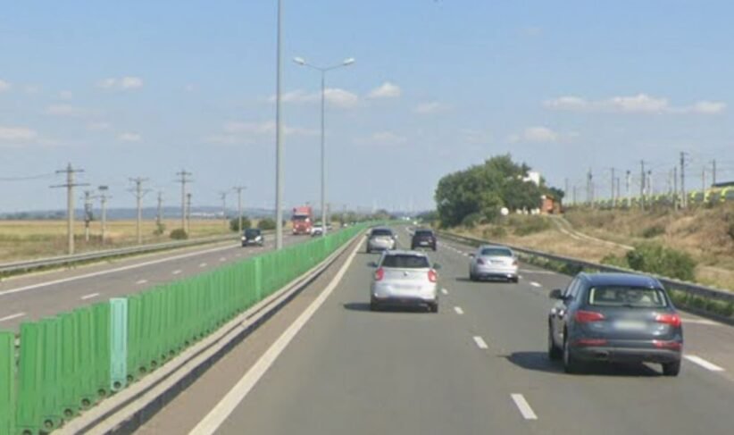 Multi-lane highway on a sunny day with green roadside barriers on the left and several cars ahead in each lane.
