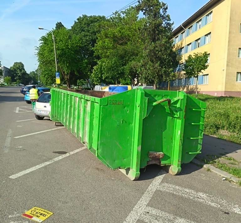 Green industrial dumpster on wheels blocking a row of parking spaces on a residential street, with trees and a yellow building in the background.