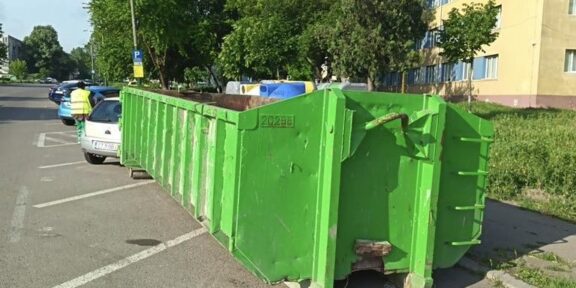 Green industrial dumpster on wheels blocking a row of parking spaces on a residential street, with trees and a yellow building in the background.