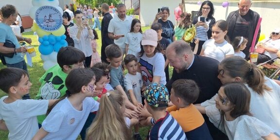 Children and adults gather in a circle with hands joined in the center at an outdoor community event, balloons in the background.