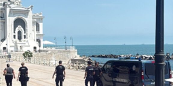 Police officers walking along a sunny seaside promenade by a white ornate building, with a dark van parked near the water.