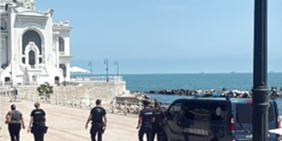 Police officers walking along a sunny seaside promenade by a white ornate building, with a dark van parked near the water.