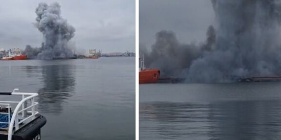 Port scene with a tall, dark smoke plume rising from a fire on the water, several ships in the distance and calm, overcast water nearby.