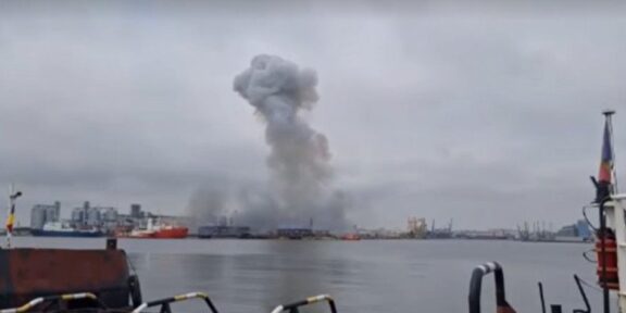 Harbor scene with a large smoke plume rising from industrial area in the distance, water in foreground and ships docked along the quay under a cloudy sky.