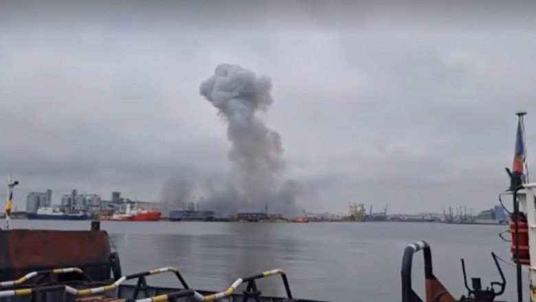 Harbor scene with a large smoke plume rising from industrial area in the distance, water in foreground and ships docked along the quay under a cloudy sky.