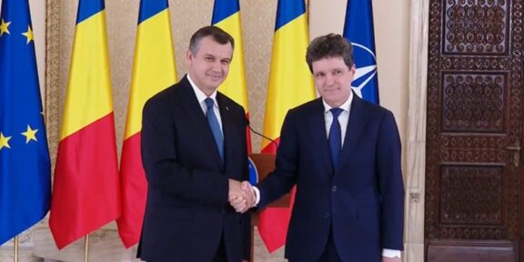 Two men in dark suits shake hands in a formal room, with EU, Romania, Ukraine, and NATO flags behind them.