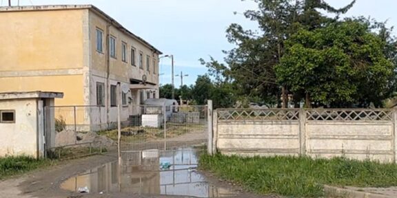 Two-story yellow building behind a chain-link gate on a muddy driveway with a large puddle and yard with trees to the right.