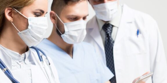 Three healthcare professionals wearing masks and white coats look at a tablet together, discussing patient care.