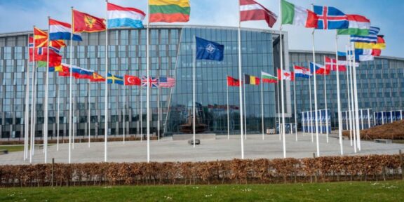 Modern glass-front building with row of flagpoles displaying numerous international flags in front of the plaza under a blue sky