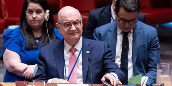 Formal meeting scene: a bald man in a navy suit and pink tie sits at a United States delegation table with microphones and documents nearby.