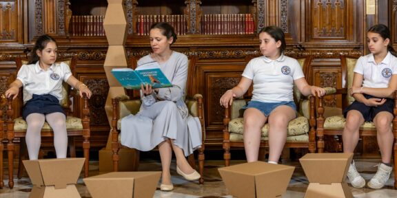 Teacher reads a blue book to four girls in white uniforms seated in an ornate wood library; cardboard art sculptures on the floor in front.