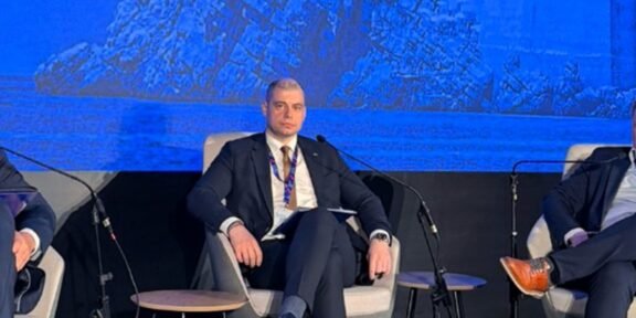 Man in a dark suit seated on a panel stage with a blue backdrop showing rocks and a coastline, microphones nearby.