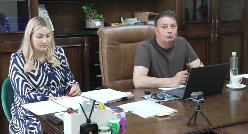 Two colleagues sit at a large wooden desk in an office, with papers and a laptop in front of them.