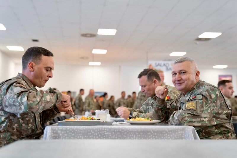 Three soldiers in camouflage sit at a table, eating in a bright mess hall with other service members in the background.